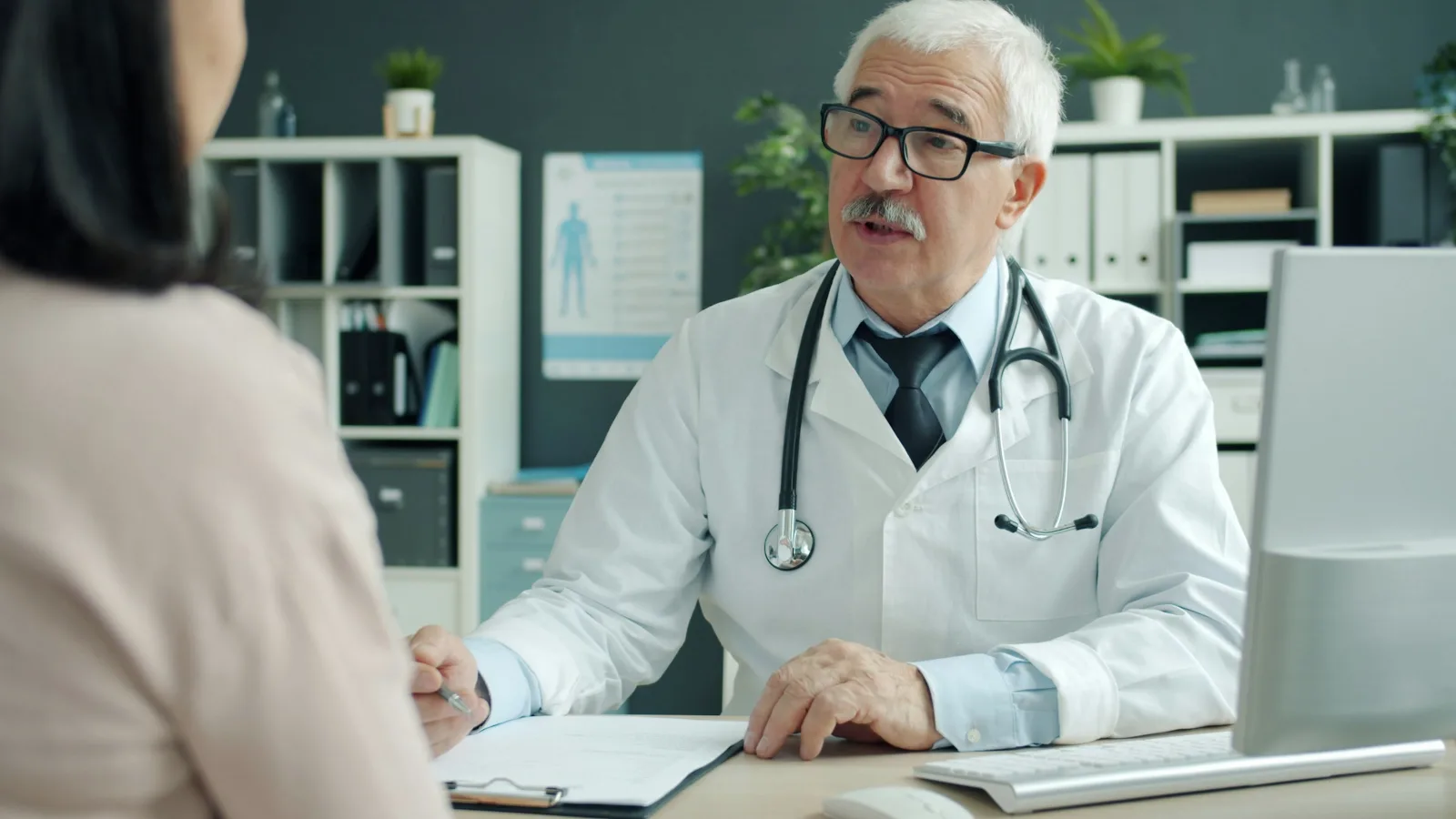 Ein Arzt spricht in seinem Sprechzimmer mit einer Patientin, die mit dem Rücken zur Kamera sitzt.