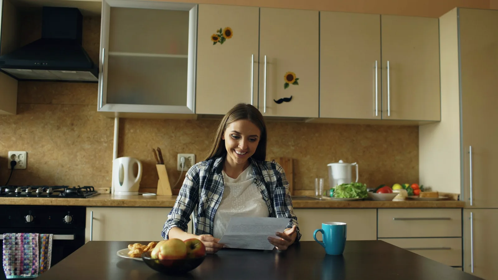 Eine Frau sitzt an einem Küchentisch und liest ein Schreiben, daneben stehen eine Tasse und eine Obstschale.